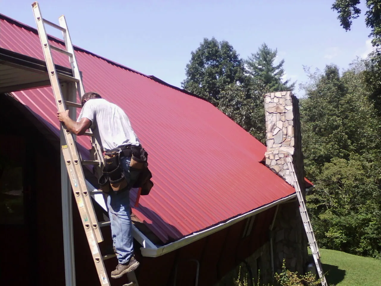 Roofer on a ladder beside a red metal roof and stone chimney — Soffit & Fascia Repair by Roofing Brothers in New Egypt